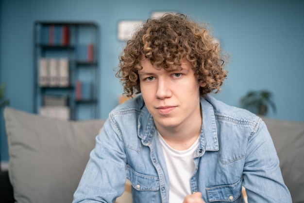 Retrato de un apuesto joven collage caucásico con cabello rizado mira a la cámara y sonríe a un hombre encantador y exitoso descansando en una sala de estar luminosa