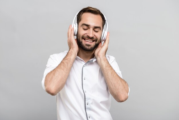 Retrato de un apuesto joven empresario con camisa blanca y corbata que se encuentran aisladas sobre una pared gris, escuchando música con auriculares inalámbricos