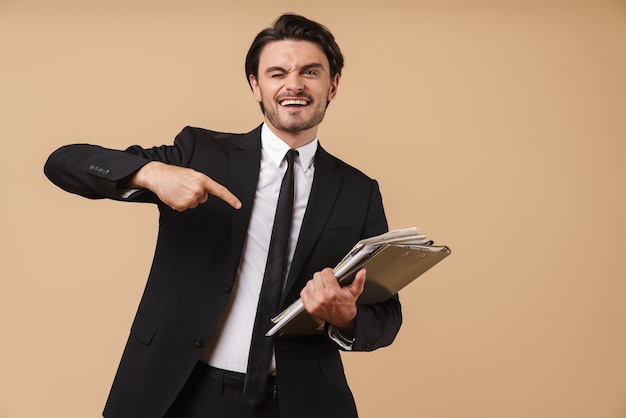 Retrato de un apuesto joven empresario confiado sonriente vistiendo traje que se encuentran aisladas sobre la pared beige, sosteniendo la pila de documentos