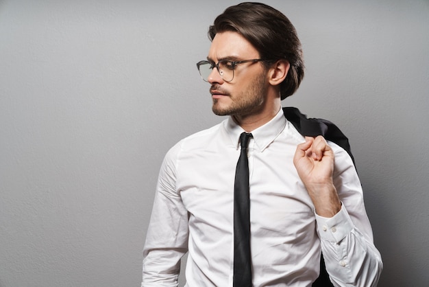 Retrato de un apuesto joven empresario confiado vistiendo traje que se encuentran aisladas sobre la pared gris, posando con chaqueta sobre su hombro