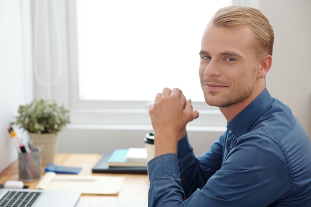 Retrato del apuesto joven empresario rubio sentado en el escritorio de oficina y sonriendo a la cámara después de terminar el trabajo en un gran proyecto