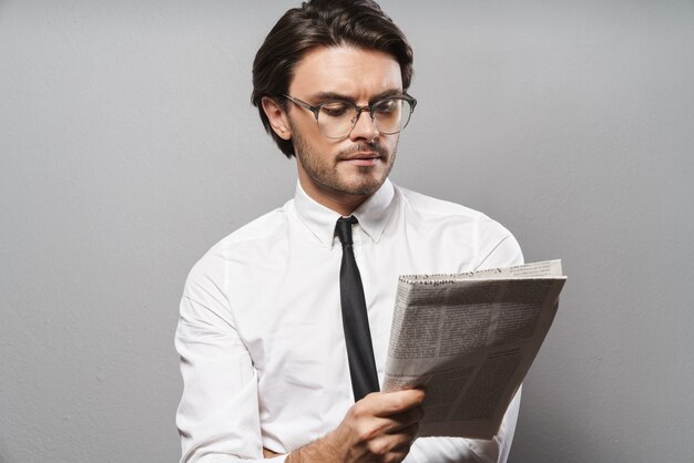 Retrato de un apuesto joven empresario serio vistiendo traje que se encuentran aisladas sobre la pared gris, leyendo el periódico