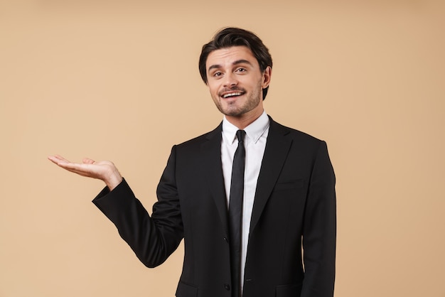 Retrato de un apuesto joven empresario sonriente vistiendo traje que se encuentran aisladas sobre pared beige, presentando espacio de copia