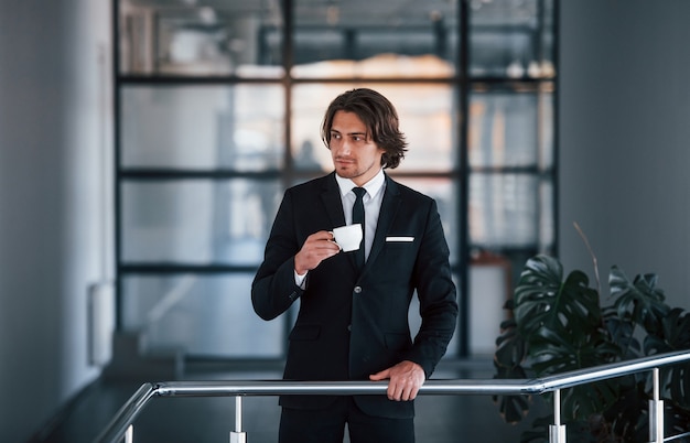 Retrato de apuesto joven empresario de traje negro y corbata se encuentra en el interior con una taza de bebida.
