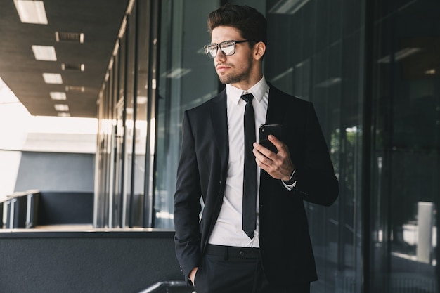 Retrato de apuesto joven empresario vestido con traje formal de pie fuera del edificio de cristal y sosteniendo el teléfono móvil