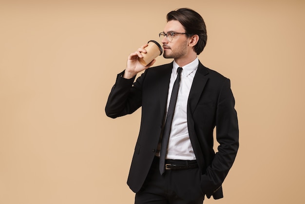 Retrato de un apuesto joven empresario vistiendo traje que se encuentran aisladas sobre pared beige, bebiendo café para llevar