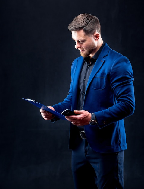 Retrato de apuesto joven exitoso vestido con camisa oscura y traje azul con carpeta azul, mirando notas. Fondo negro. Concepto de negocio.