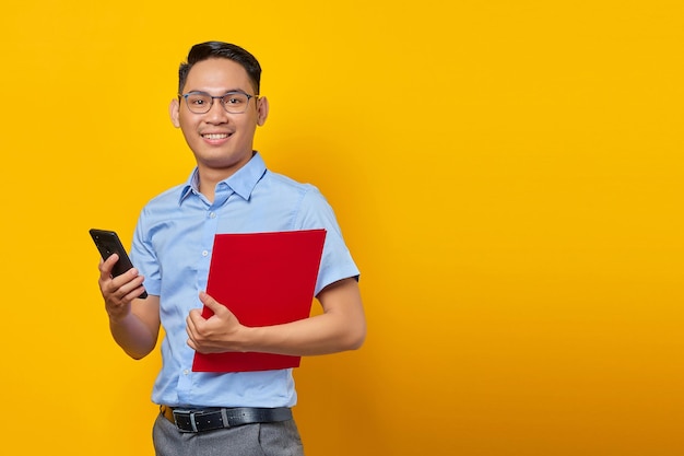 Retrato de un apuesto joven hombre de negocios asiático con anteojos de pie sosteniendo con confianza una carpeta de documentos y usando un teléfono móvil con cara sonriente aislada sobre fondo amarillo