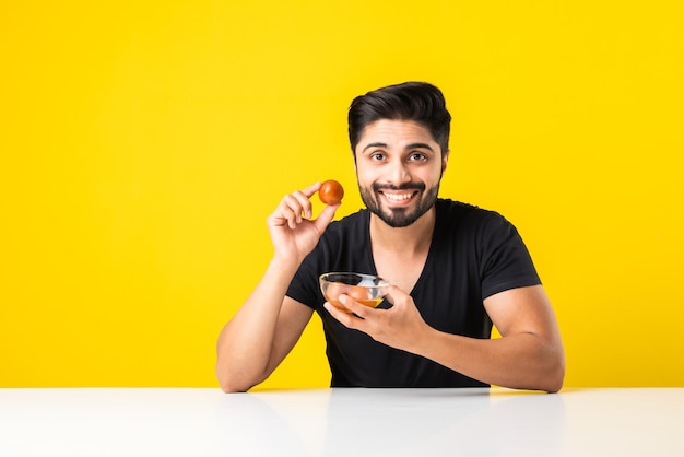 Retrato de un apuesto joven indio manÃ'Â comiendo dulce Gulab Jamun contra el fondo amarillo