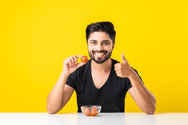 Retrato de un apuesto joven indio manÃ'Â comiendo dulce Gulab Jamun contra el fondo amarillo