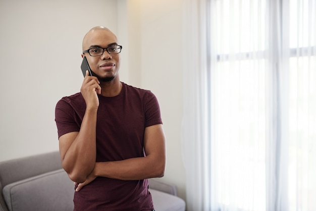 Retrato de apuesto joven negro de pie en la sala de estar y hablando por teléfono con un amigo o colega