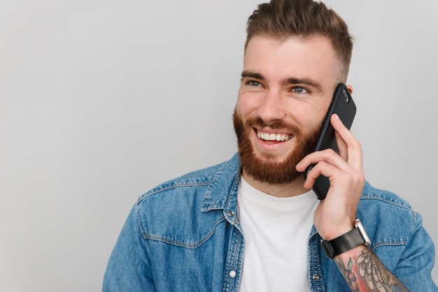 Retrato de un apuesto joven sonriente vistiendo ropa casual que se encuentran aisladas sobre la pared gris, hablando por teléfono móvil