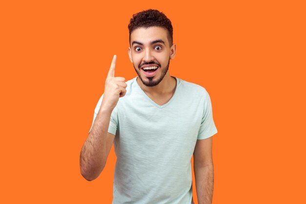 Retrato de un apuesto moreno inspirado con barba en una camiseta blanca que señala con el dedo hacia arriba y se ve asombrado por la repentina idea genial, tiene una solución. tiro de estudio interior aislado sobre fondo naranja