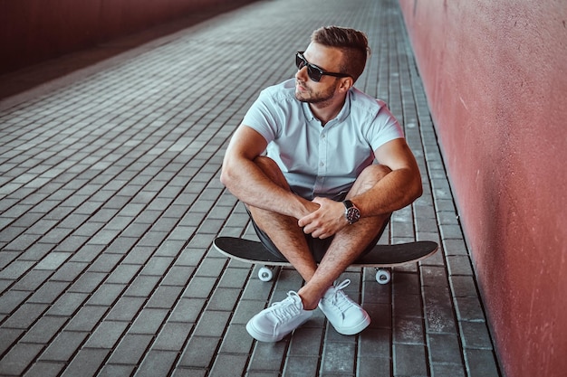 Retrato de un apuesto patinador de moda con gafas de sol vestido con una camisa blanca y pantalones cortos sentados en una patineta debajo del puente, mirando hacia otro lado.