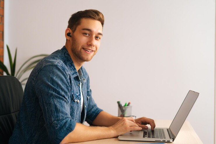 Foto retrato de un apuesto programador independiente sonriente con auriculares inalámbricos trabajando en una computadora portátil sentado en el escritorio de la oficina en casa