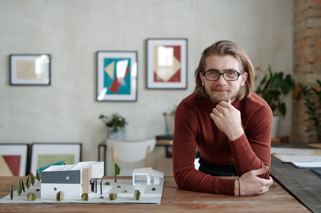 Retrato del arquitecto caucásico joven sonriente en anteojos apoyado en la mesa con el modelo 3D del edificio contemporáneo en la oficina