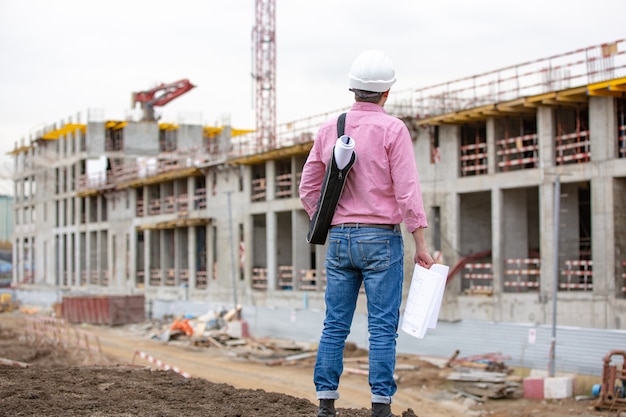 Retrato del arquitecto en el trabajo con casco en un sitio de construcción, lee el plan, proyectos en papel