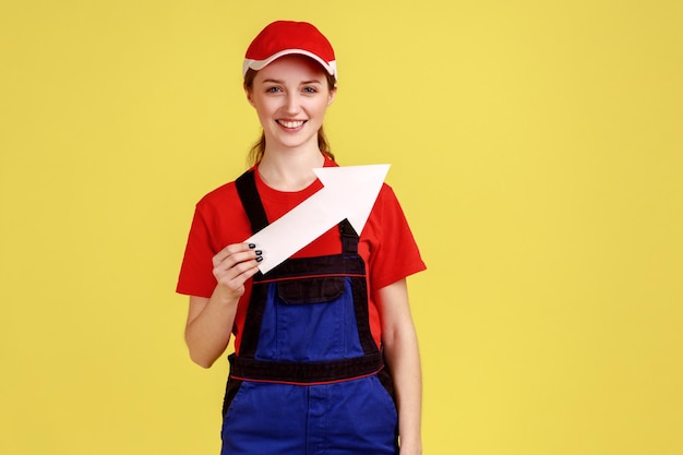 Retrato de una artesana positiva sonriente de pie con una flecha blanca en las manos que indica a un lado mirando a la cámara con un mono y una gorra roja Disparo de estudio interior aislado sobre fondo amarillo