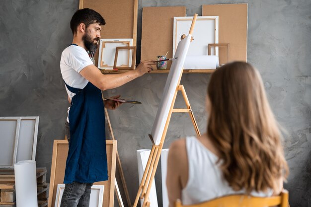 Retrato de un artista barbudo en un estudio con un delantal sucio junto al lienzo en un caballete sosteniendo un pincel y una paleta con pinturas pinta un retrato de una modelo femenina