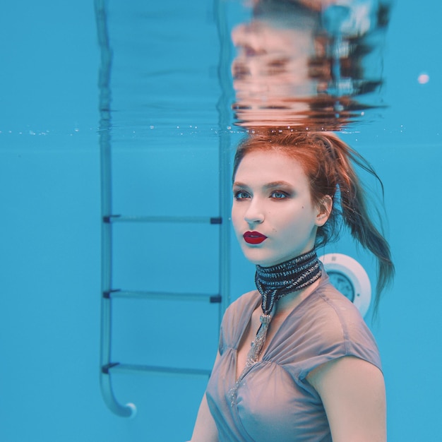 retrato artístico surrealista de una joven vestida de gris y bufanda con cuentas bajo el agua en la piscina