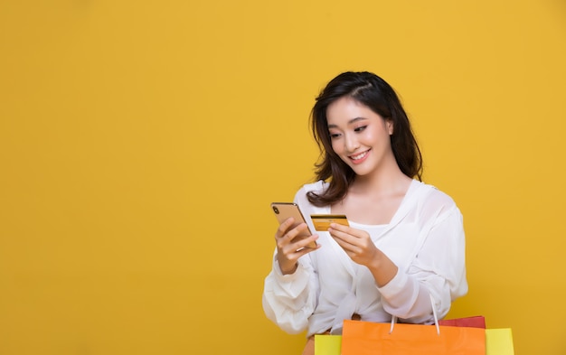 Retrato asiática feliz mulher jovem e bonita sorrindo alegre e ela está segurando o cartão de crédito e usando telefone inteligente para compras on-line com sacos de compras em fundo amarelo.