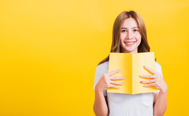 Retrato asiática tailandesa hermosa joven feliz estilo de vida mujer sostiene un libro amarillo o un diario mirando a la cámara, foto de estudio aislada en fondo amarillo, con espacio para copiar