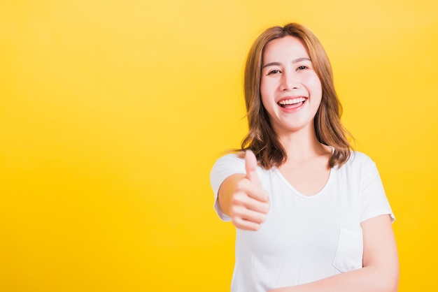 Retrato asiática tailandesa hermosa mujer joven feliz sonriendo usar camiseta blanca de pie mujer exitosa dando un pulgar hacia arriba gesto gesto en foto de estudio, aislado en fondo amarillo con espacio de copia