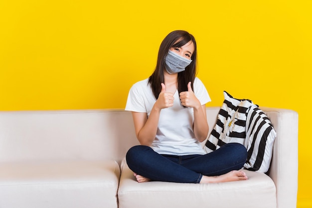 Retrato asiático de uma bela jovem sentada no sofá usando máscara médica protetora mostra o dedo polegar para cima para um bom sinal durante o tiro de estúdio do Coronavirus isolado em fundo amarelo