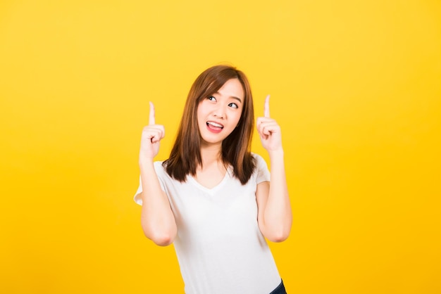 Retrato asiático feliz hermosa linda joven adolescente de pie usando una camiseta hace un gesto con dos dedos apuntando hacia arriba mirando a la cámara aislada, tiro de estudio sobre fondo amarillo con espacio de copia