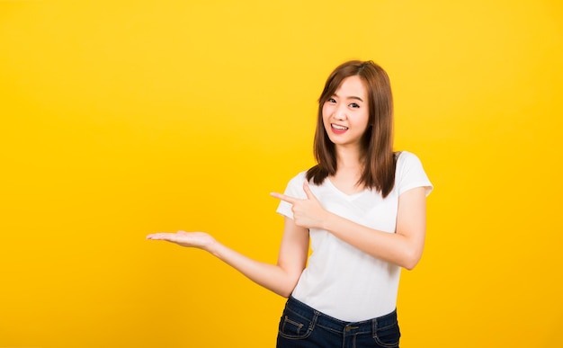 Retrato asiático feliz hermosa linda joven adolescente de pie usando una camiseta sosteniendo algo en la palma y apuntando hacia el lado mirando a la cámara aislada, tiro del estudio sobre fondo amarillo con espacio de copia