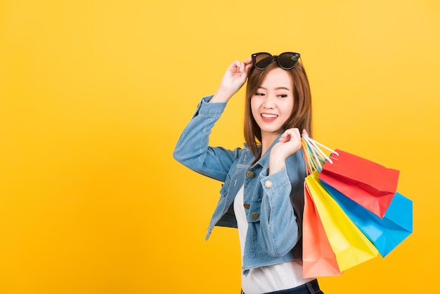 Retrato asiático feliz hermosa linda mujer joven adolescente sonriendo de pie con gafas de sol emocionado sosteniendo bolsas de compras bolsas de aspecto multicolor aisladas, estudio de fondo amarillo con espacio de copia