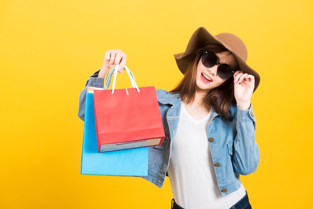 Retrato asiático feliz hermosa linda mujer joven adolescente sonriendo de pie con gafas de sol emocionado sosteniendo bolsas de compras cámara de aspecto multicolor aislada, estudio de fondo amarillo con espacio de copia