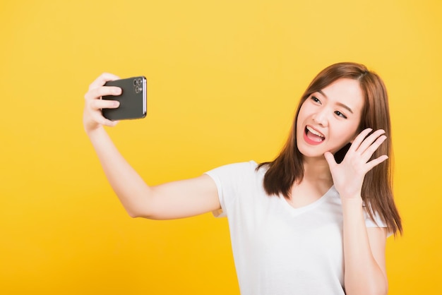 Retrato asiático feliz hermosa linda mujer joven adolescente sonriendo de pie haciendo una foto selfie, videollamada en el teléfono inteligente levanta la mano para decir hola aislado, estudio de fondo amarillo con espacio de copia