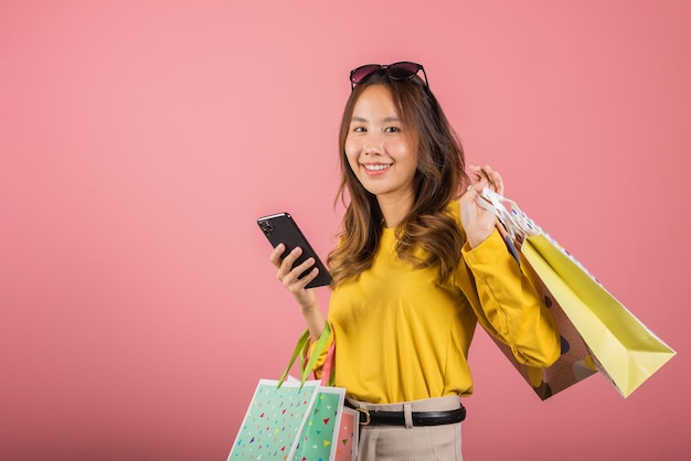 Retrato asiático feliz hermosa mujer joven adolescente comprador sonriente de pie emocionado sosteniendo bolsas de compras en línea en línea multicolor en teléfono móvil en la mano tiro de estudio aislado