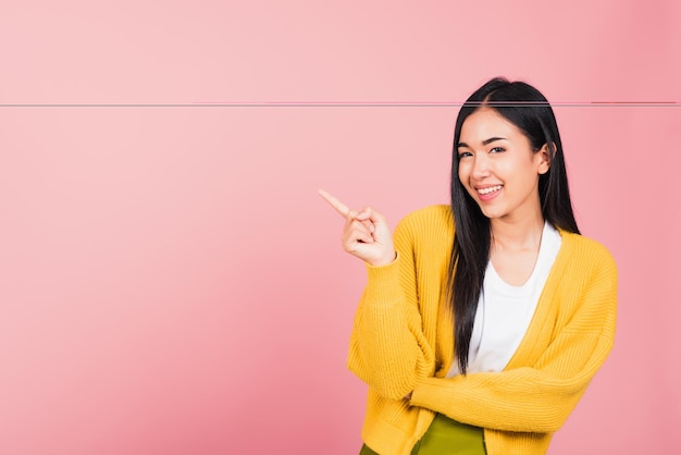 Retrato asiático feliz hermosa mujer joven linda de pie apuntando con el dedo al lado presentando el producto mirando a la cámara, foto de estudio aislada en fondo rosa con espacio de copia