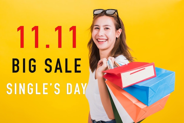 Retrato asiático feliz hermosa mujer joven sonriendo de pie con gafas de sol emocionado sosteniendo bolsas de compras multicolor con 11.11 gran venta y texto del día del soltero en el espacio aislado fondo amarillo