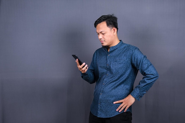 Retrato de un asiático infeliz con camisa azul sosteniendo un teléfono móvil con expresión triste en la cara.