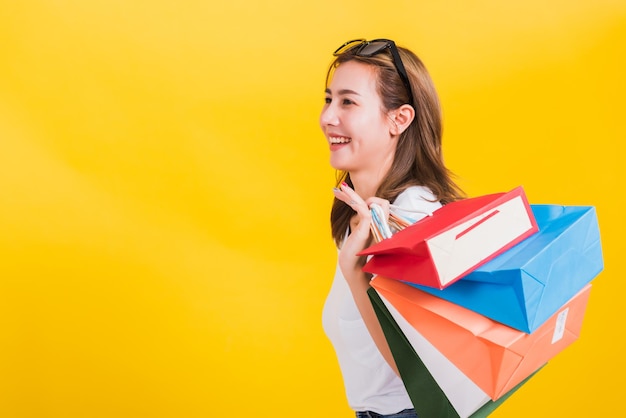 Retrato asiático tailandés feliz hermosa linda mujer joven sonriente de pie con gafas de sol emocionada sosteniendo bolsas de compras de varios colores mirando al lado, foto de estudio aislado fondo amarillo con espacio de copia