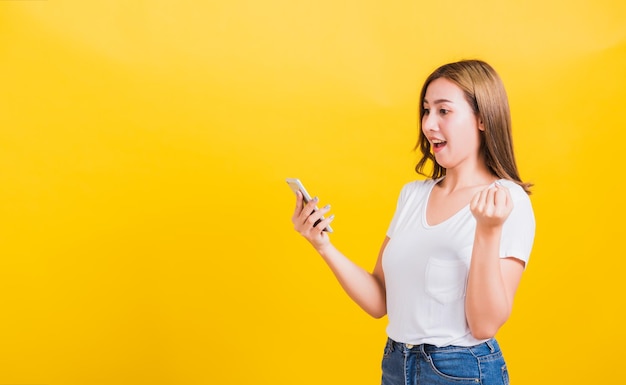 Retrato asiático tailandês feliz linda jovem bonita sorridente vestindo camiseta levante mão comemorando a vitória com telefone celular inteligente olhando o telefone isolado, em fundo amarelo com espaço de cópia