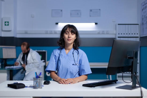 Retrato de asistente médico usando la computadora en el escritorio para trabajar horas extras. Enfermera de la mujer en uniforme que mira la cámara preparándose para el papeleo y el chequeo médico, trabajando hasta tarde en la oficina.