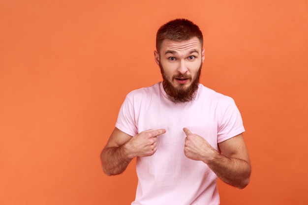 Retrato de un asombrado y apuesto hombre barbudo apuntándose a sí mismo, mirando a la cámara con grandes ojos sorprendidos, usando una camiseta rosa. Disparo de estudio interior aislado sobre fondo naranja.