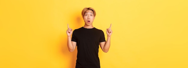 Retrato de asombrado chico asiático en camiseta negra boca abierta fascinado señalando con el dedo hacia arriba amarillo ba