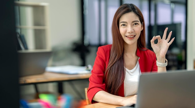 Retrato de una atractiva y alegre chica habilidosa con traje rojo que usa una computadora portátil y muestra el signo de aprobación mirando la cámara y sentada en el escritorio