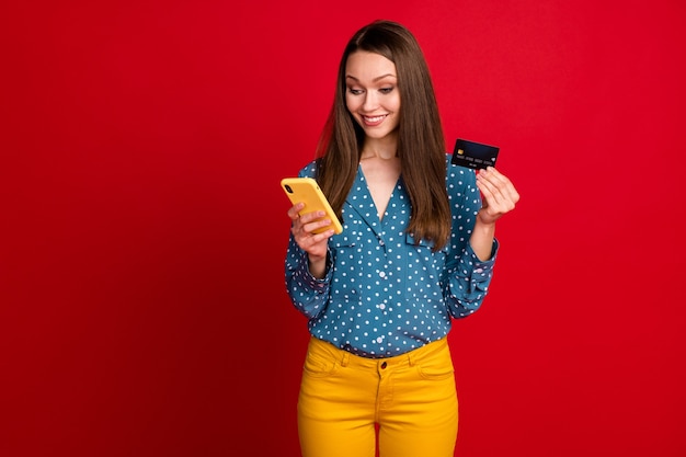 Retrato de atractiva chica alegre enfocada usando la aplicación de servicio de pago de tarjeta bancaria de dispositivo aislada sobre fondo de color rojo brillante