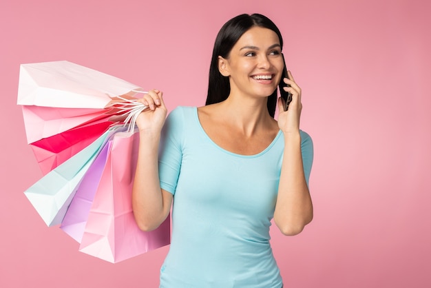 Retrato de atractiva chica alegre de pelo lacio sosteniendo en la mano llevando cosas nuevas y conversando en el teléfono aislado sobre fondo de color rosa brillante