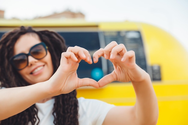 Retrato de una atractiva chica alegre con rastas onduladas divirtiéndose al aire libre mostrando un signo de corazón afuera