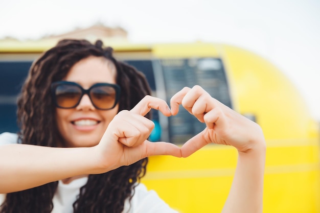 Retrato de una atractiva chica alegre con rastas onduladas divirtiéndose al aire libre mostrando un signo de corazón afuera