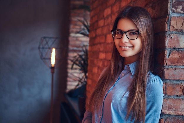 Retrato de una atractiva y encantadora morena con gafas y camisa azul sonriendo y apoyada en una pared de ladrillos en una habitación con diseño de loft.