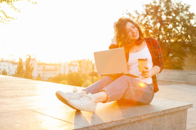 Retrato de una atractiva estudiante sonriente. Joven hipster usa laptop y sostiene una taza de café en la ciudad al amanecer