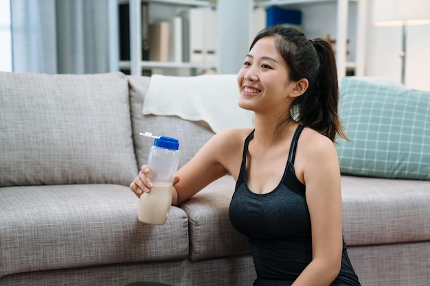 retrato de una atractiva y hermosa mujer china asiática en ropa deportiva con una botella de leche apoyada en el sofá en la sala de estar de casa. vista lateral joven sonriente disfruta de una bebida proteica después del yoga.